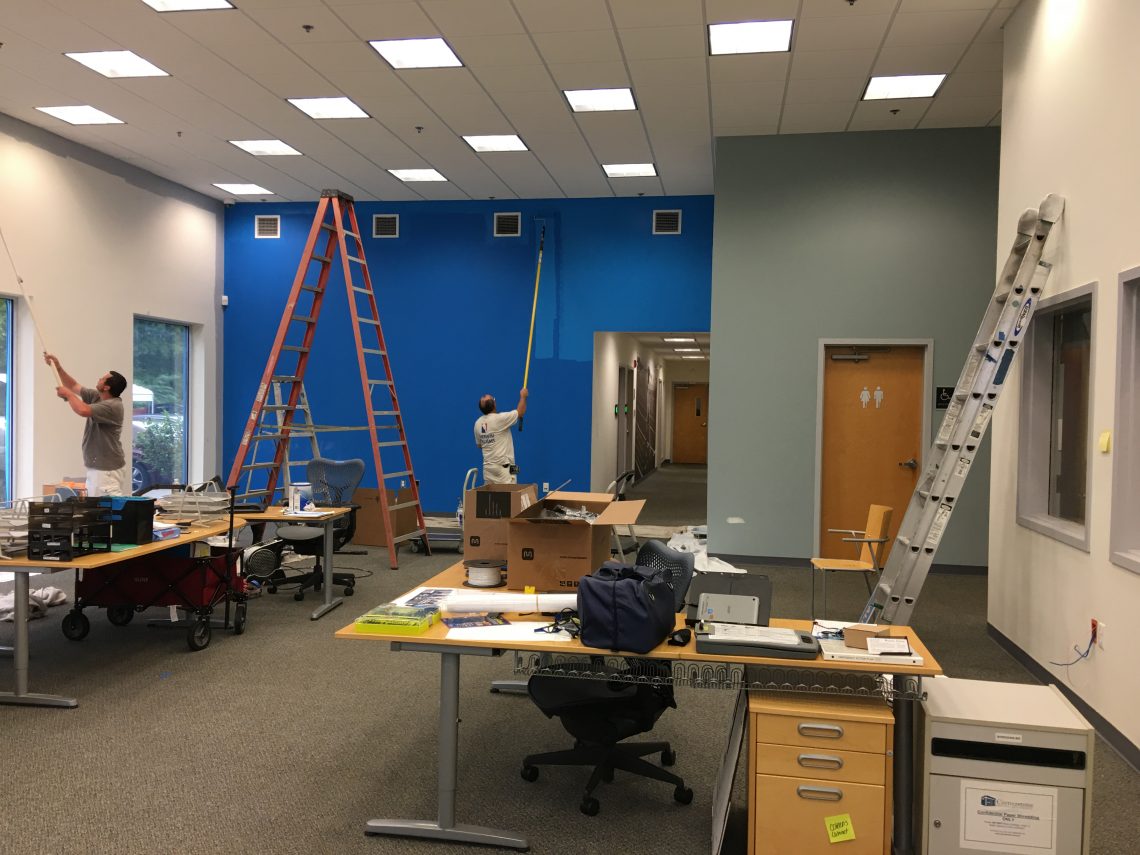 Interior commercial office space being painted by two men using rollers and long poles