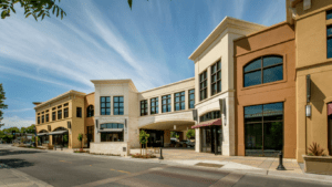 The exterior of a retail shopping center with stucco siding painted tan, gold, brown, and white. There is an open breezeway in the middle leading to parking and a street occupies the foreground.