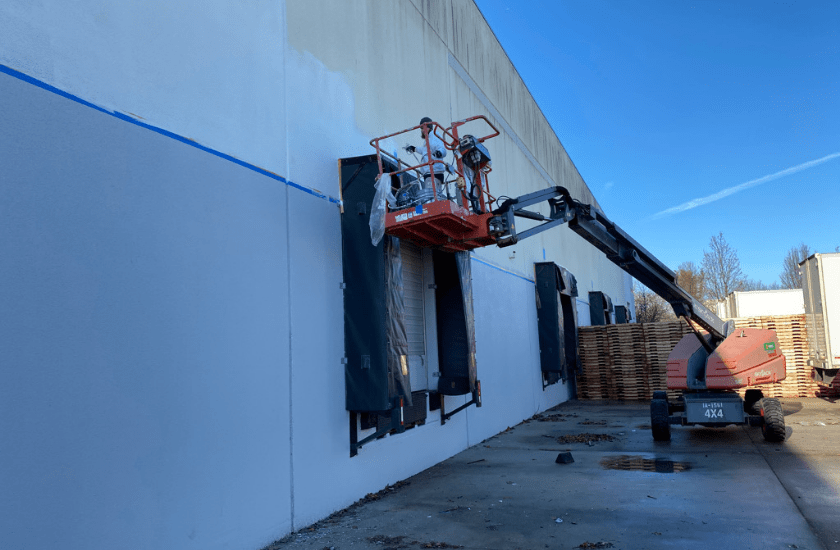 A painter from DJ's Painting is using a red and black boom lift to paint above an overhead door on the exterior of a warehouse with poured concrete walls.The upper portion of the building is being painted white and the bottom has been painted gray.